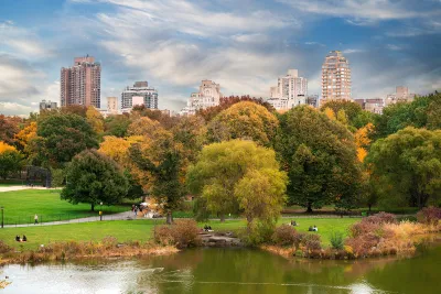 View of green meadow with people and fall-colored trees in Central Park with Manhattan buildings in background.