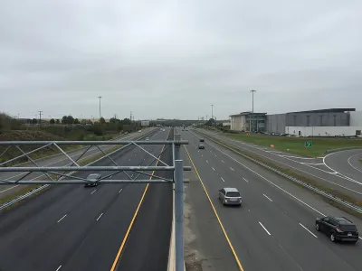 A wide highway with a few cars on a gloomy day. Low-rise commercial buildings and more car infrastructure is visible to the side of the road.