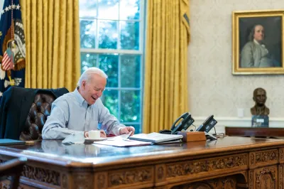 President Jose Biden appears in a collared, button down shirt while signing papers while seated at the desk in the Oval Office of the White House
