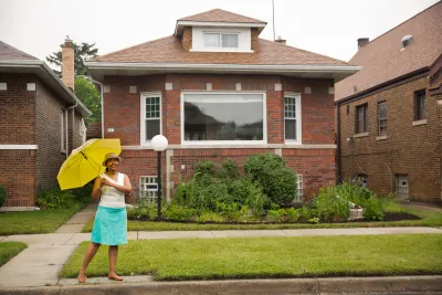A Black woman stands in front of a home holding an umbrella and smiling.