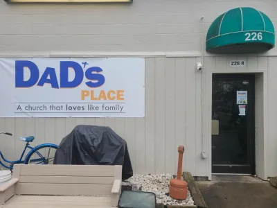 White sign on side of tan building with metal siding and green overhang, reading "Dad's Place, a church that loves like family."