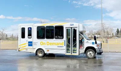 White minibus with yellow "On-Demand" logo parked in an asphalt lot with grass field and dry fall trees in background in Edmonton, Canada.