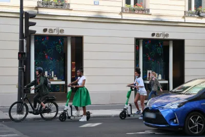 A woman on an electric bike and two women on electric scooters wait at an intersection for the light to change.