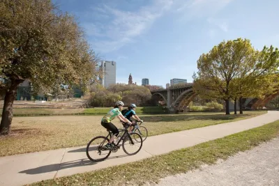 Two cyclists on a paved bike trail through short grass with Dallas buildings in background