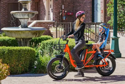Woman in purple helmet on orange e-bike with baby in seat on back of bike.