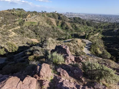 The Griffith Observatory is perched on a hill above Los Angeles, with the city's downtown shown int he distant background. Griffith Park's rolling hills and winding roads are shown in the foreground.