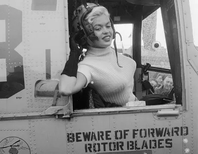 Black and white photo of actress Jayne Mansfield leaning out of the window of a military plane wearing flight helmet for promotional photo