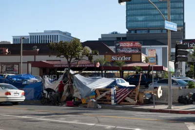A homeless encampment fills a sidewalk in front of a Sizzler restaurant advertising Thanksgiving dinner.