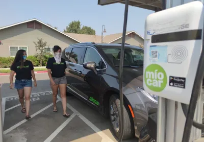 Black SUV charging at Miocar electric car sharing station with two women walking towards it.