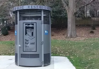 Portland Loo modular gray public bathroom booth sits on concrete slab with green lawn and tree in background in a park in Portland, Oregon
