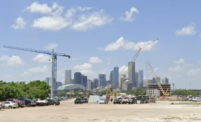 Dirt lot with construction cranes and parked cars with downtown Houston skyline in background