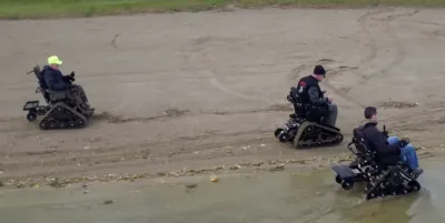 Aerial view of three men using track chair mobility devices on lake beach in Michigan state park.