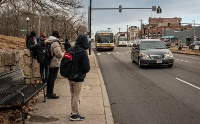 People in hoodies and jackets waiting for oncoming bus at sidewalk bus stop in Boston, Massachusetts.