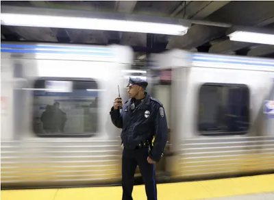 Transit police officer standing on subway platform with blurred train passing behind him.