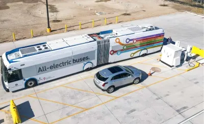Aerial view of electric bus parked next to silver SUV in parking lot in Austin, Texas. Bus says "All eelctric buses" on side.