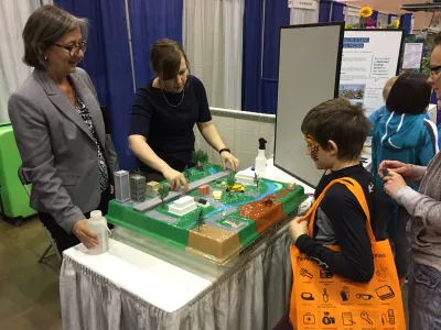 Two adults stand behind a table, gesturing toward a model of a city while talking to a young child about buildings and parks and streets.