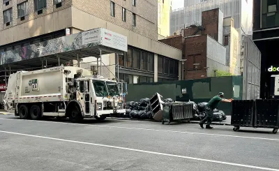 White waste management sanitation garbage truck and worker pushing wheeled green dumpster on street with piles of trash bags.