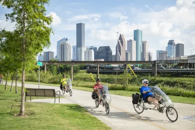 Three men riding recumbent bikes on paved greenway next to bayou with Houston skyline in background.