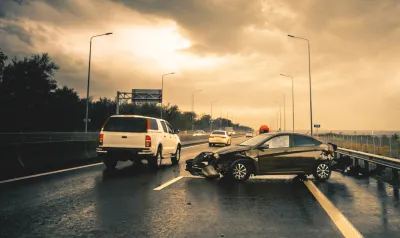Small black car with mangled bumper sitting sideways on an asphalt road