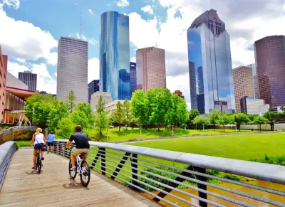 Two cyclists on a wooden bike bridge across a bayou with downtown Houston skyline in background