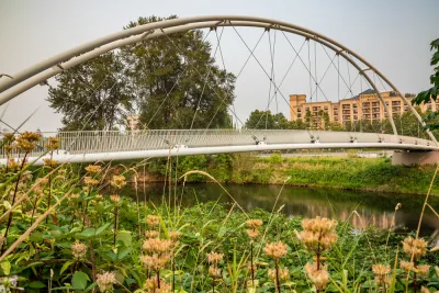 View of pedestrian suspension bridge over waterway surrounded by green grass with small apartment buildings in background
