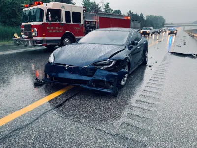 Dark blue Tesla with crumpled front bumper on rainy highway with red fire truck in background