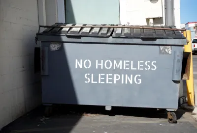 "No homeless sleeping" stenciled on grey dumpster in urban alley