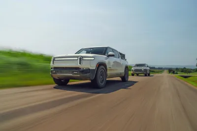 Two silver Rivian electric pick-up trucks driving down road with blurry grass on roadside