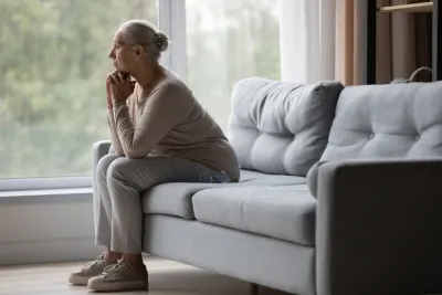 Side view of older woman sitting on gray sofa gazing pensively out of wall-sized window.