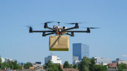 Delivery drone holding a brown paper wrapped box hoveringin air with city in background.