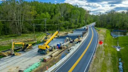 Aerial view of road construction with yellow heavy equipment parked next to two-lane asphalt road against green forest and blue sky