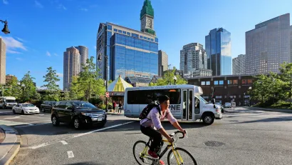 Person on bike wearing backpack with small bus and Boston buildings in background