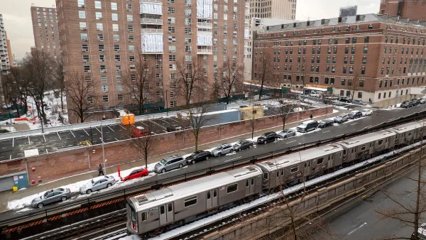 Silver New York City subway train on above ground track with red brick multistory buildings in background.