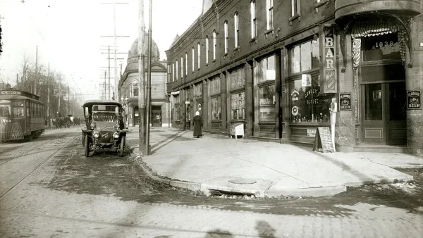 Black-and-white photo of street with old black model T and brick building on the corner.
