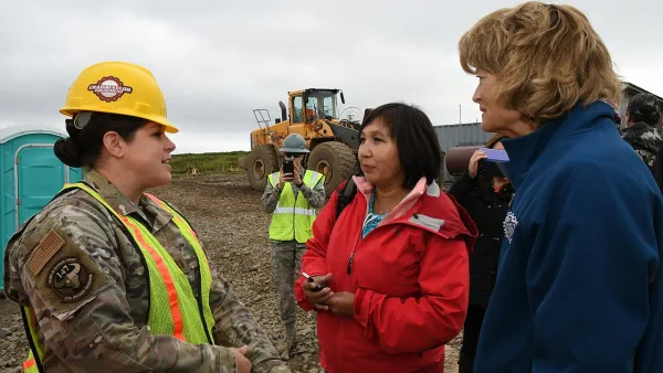 Three people, one in fatigues and hard hat, standing in front of bulldozer discussing relocation plans in Newtok, Alaska.
