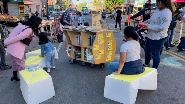 Children sit on temporary street furniture next to book cart at pop-up reading event at open streets event in Sunset Park in New York City.