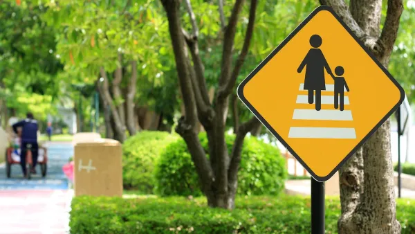 Yellow pedestrian crossing sign with black stick figures of mother and child crossing white crosswalk with blurred green trees on city street in background.