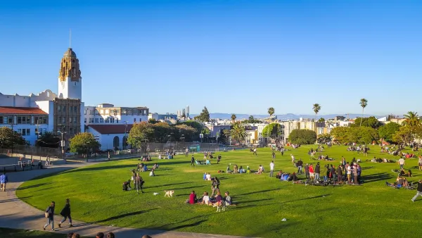 View of people sitting in lightly sloped green lawn with Spanish colonial-style building and cityscape in background in San Francisco, California.