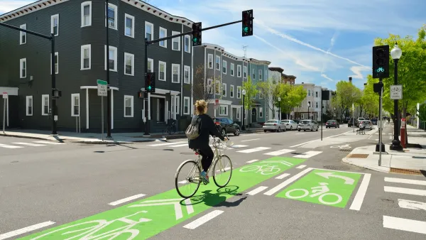 Cyclist on bike in green painted bike lane at intersection with three-story buildings in background.