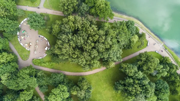Bird's eye view of park with green trees, concrete walkways, and benches on lake or riverfront.