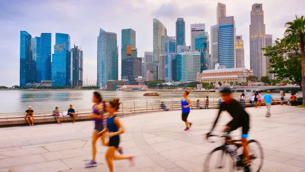 People running and cycling on wide paved path next to water with Singapore skyline with high-rises in background.