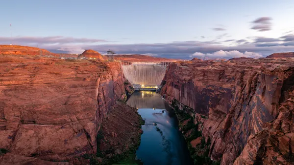 View of Glen Canyon Dam with vertical red stone cliffs on either side, Colorado River at bottom, and dusk sky in background.