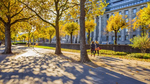 Two women walking on wide walkway among large trees next to neoclassical building in Cologne, Germany.