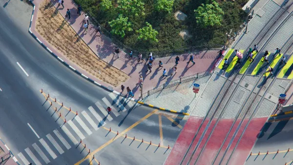 Overhead view of doxens of commuters walking on sidewalk and crossing street in full sun.