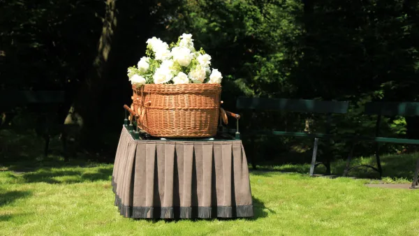 Woven willow casket with white flowers on top sits on table with pleated tablecloth on green lawn