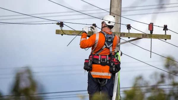 Worker in orange shirt and white helmet works at top of power line.