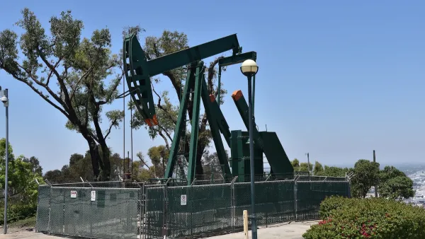 Green oil well pump jack surrounded by fence on a hill overlooking neighborhood.