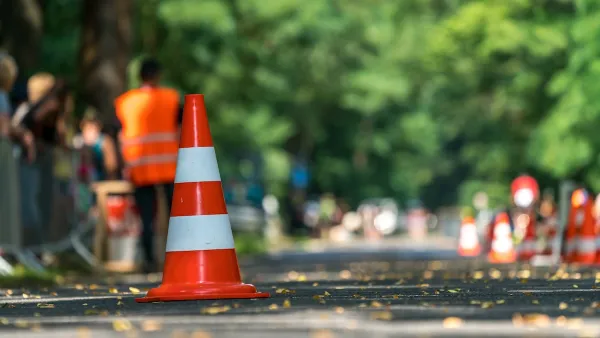 Close-up of orange and white traffic cone on street with blurred worker in orange vest in background.
