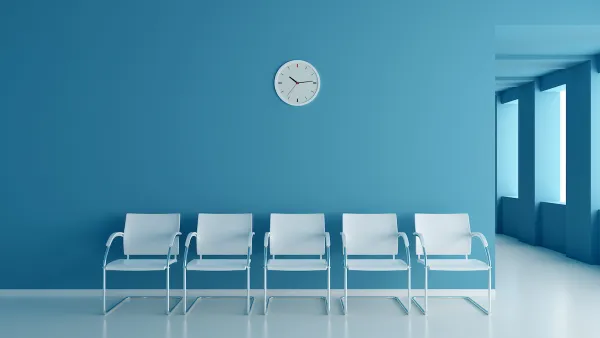 Row of five white chairs set against a blue wall with a round white analog clock. Clinic waiting room.