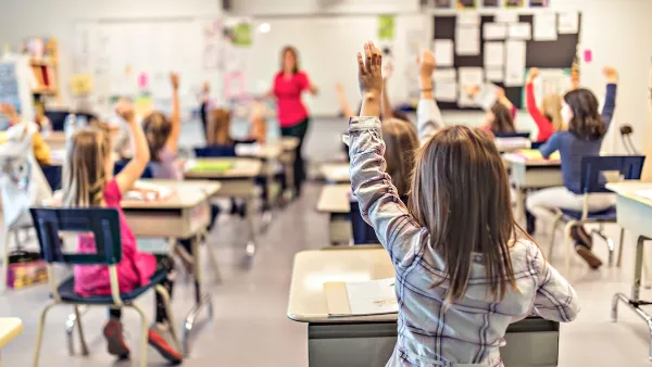 View from back of classroom of elementary school children at desks with raised hands engaged in class.